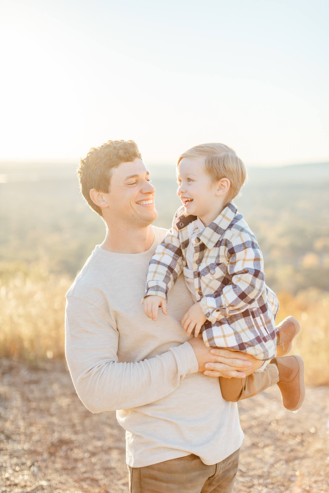 A happy toddler plays in dad's arms in the hills at sunset in plaid and tan