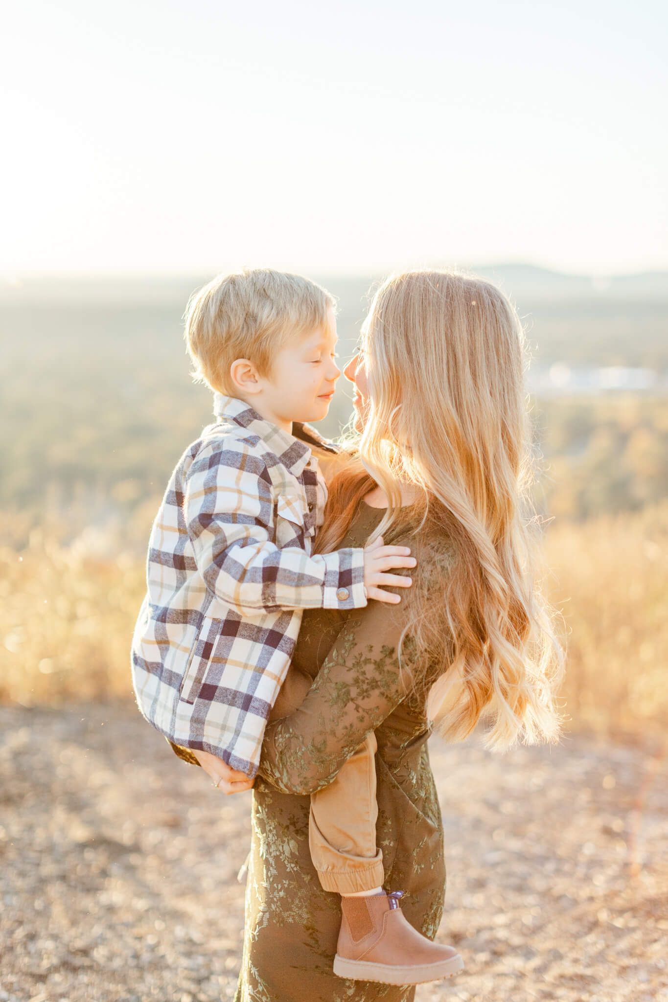 A happy mom in a lace dress boops noses with her toddler son in plaid sitting in her arms on an overlook at sunset after exploring preschools in huntsville al