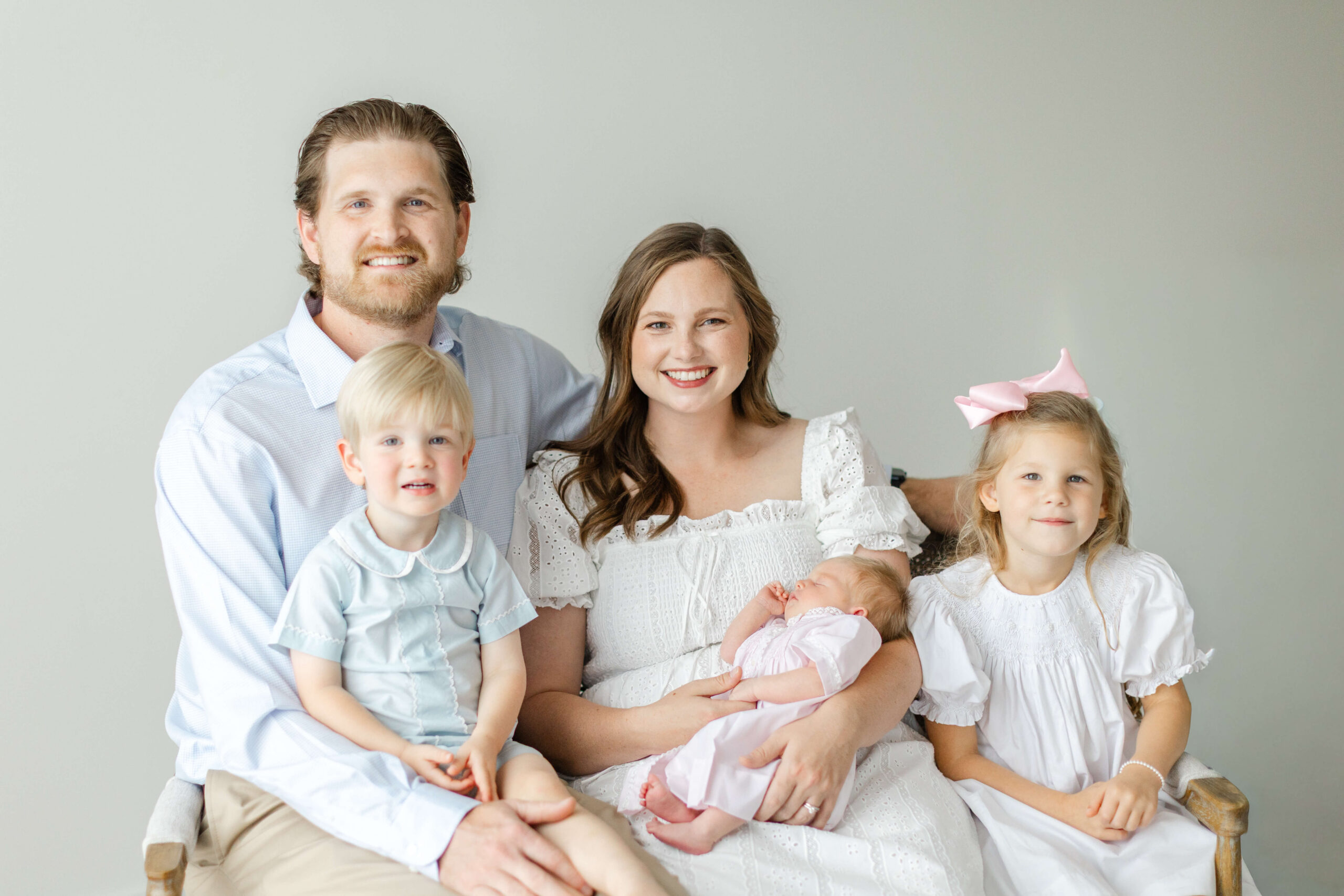 A smiling mom and dad sit on a bench with their two toddlers and newborn all in blue and white after mom looked into The Bunny Hive Huntsville