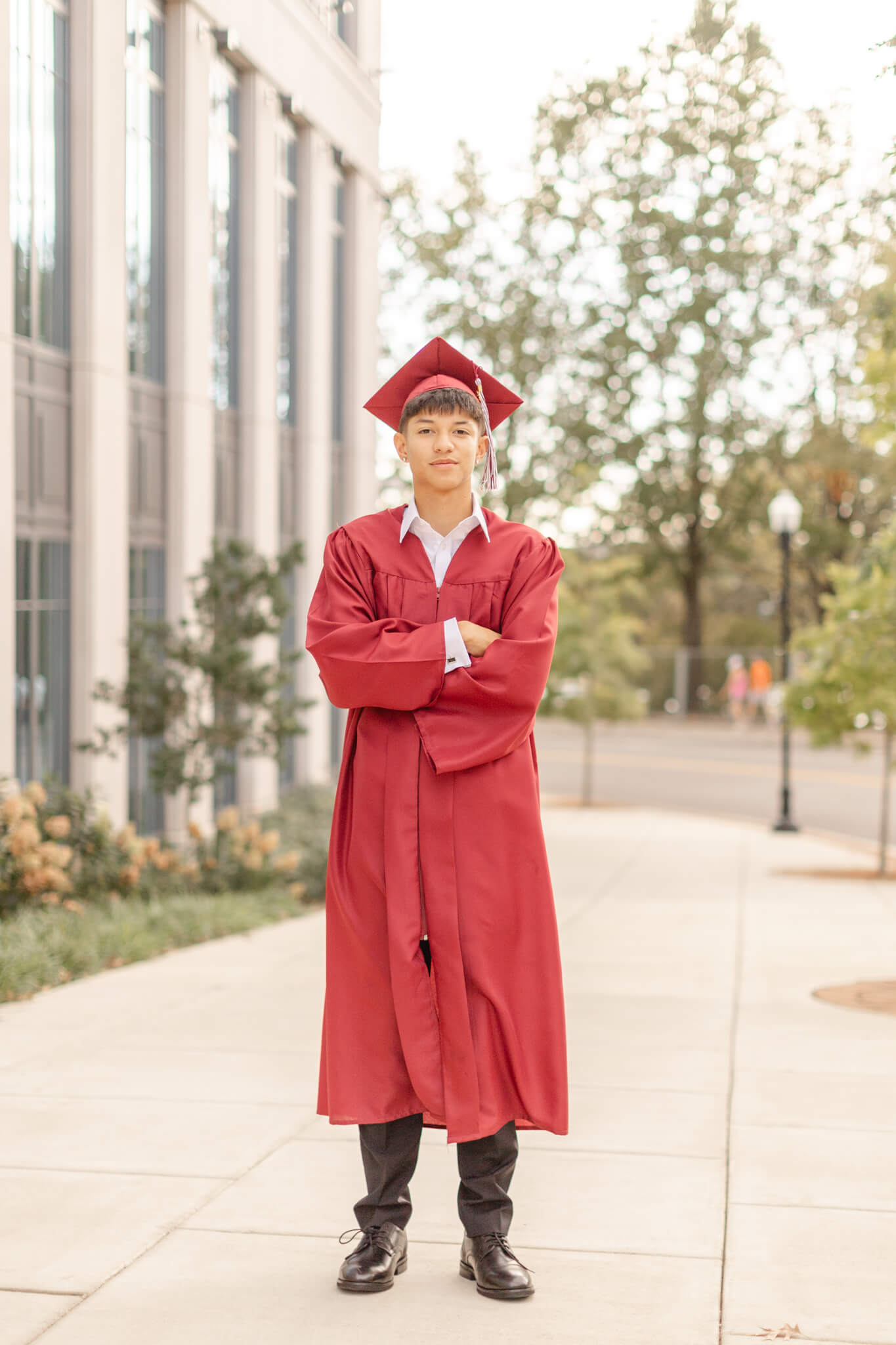 A high school senior in his red cap and gown stands with arms crossed on a sidewalk after looking into colleges near huntsville al