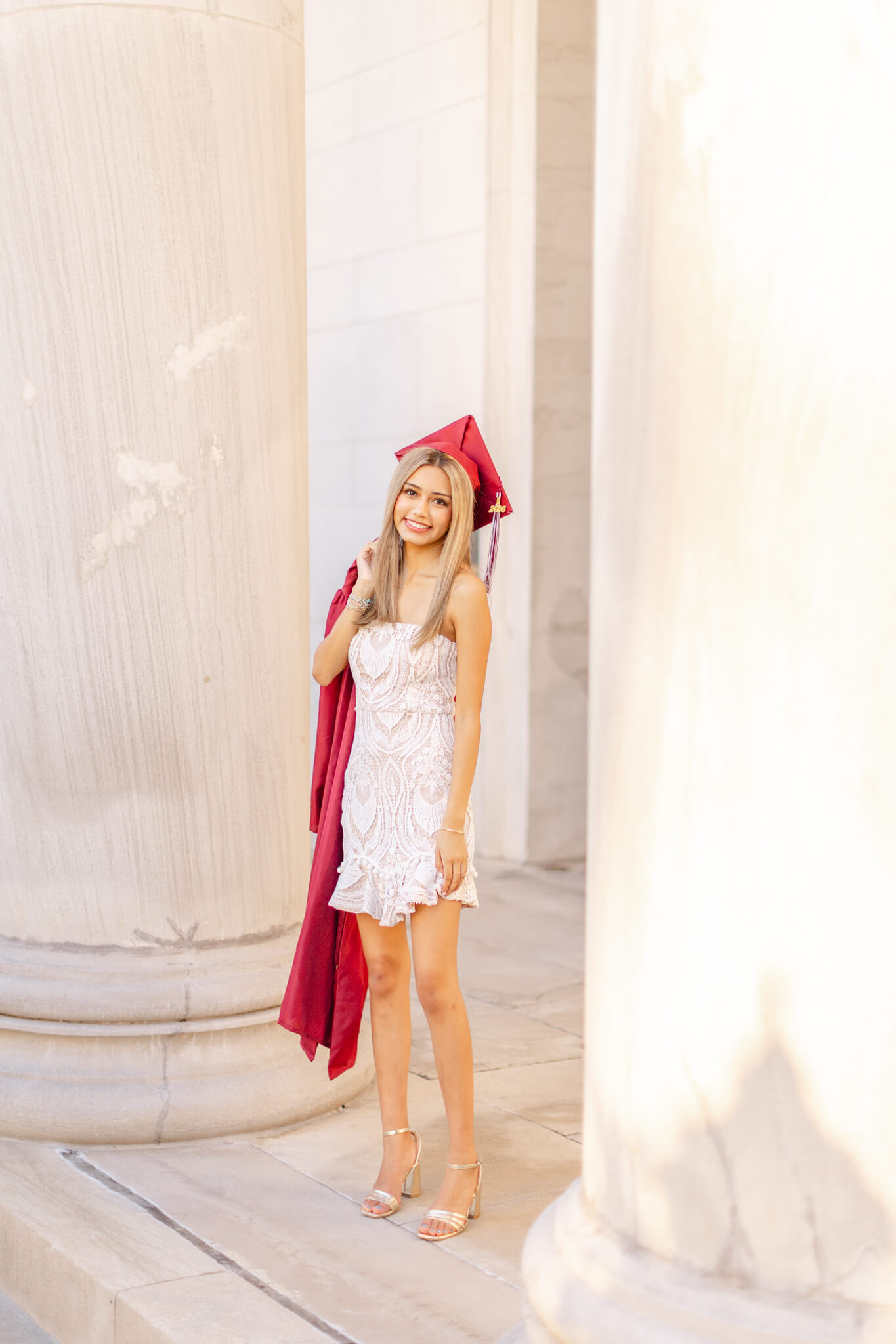 A woman in a white dress stands between two large columns with her red gown over her shoulder