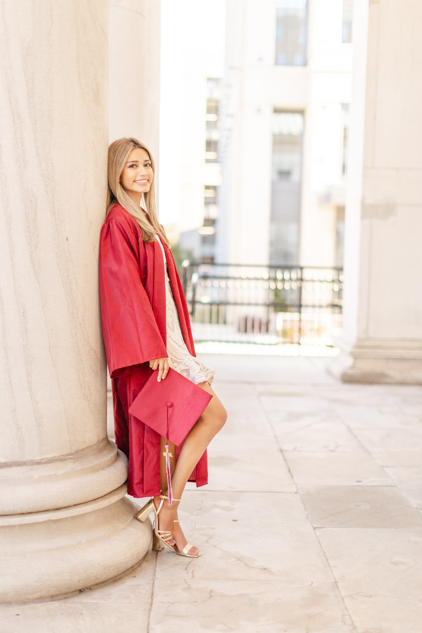 A graduate in a red gown leans against a large column smiling after finding graduation party venues in huntsville al