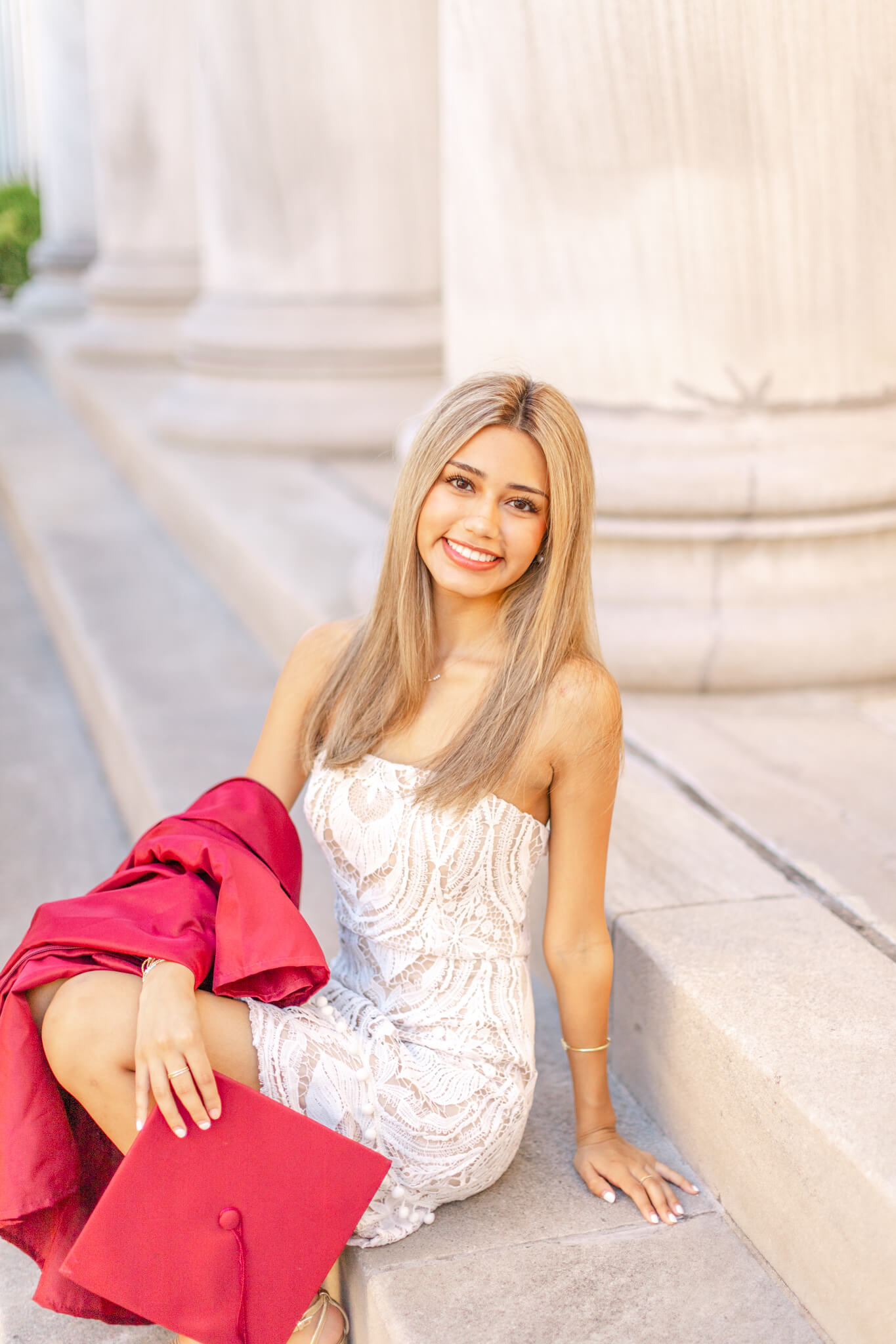 A smiling grad sits on granite steps in a white dress with red cap and gown in her lap after exploring graduation party venues in huntsville al