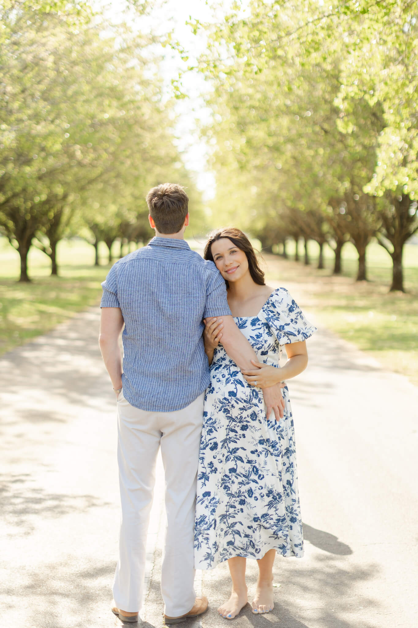 A happy mother to be leans on the shoulder of her husband in a blue and white floral print maternity dress in a park path lined with trees at sunset