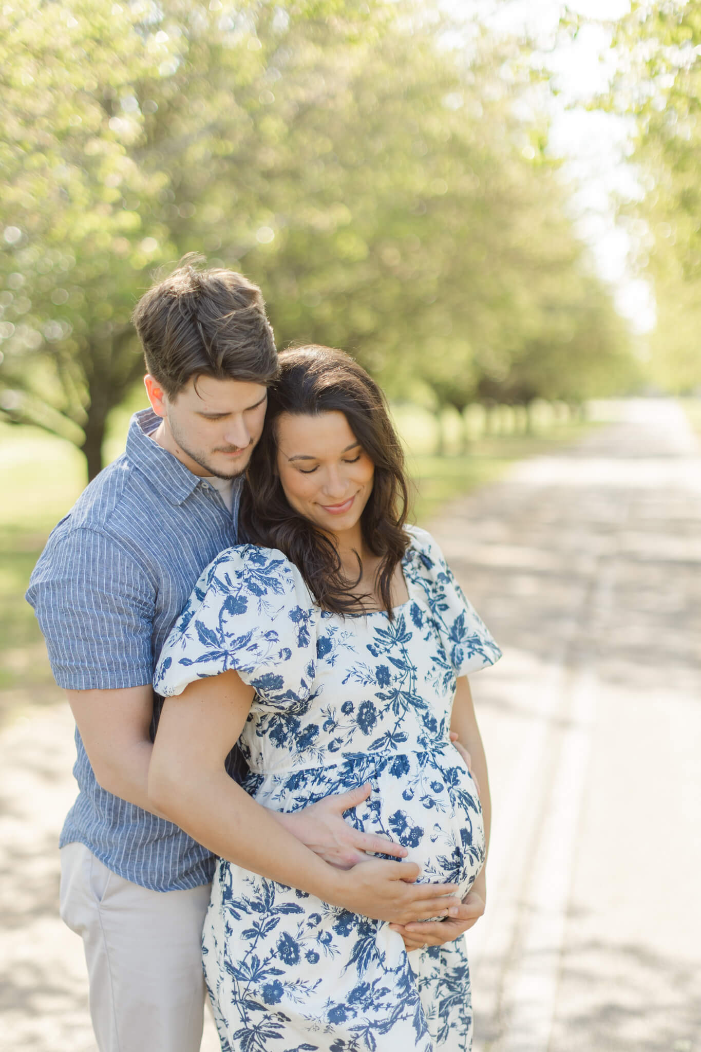 Parents to be smile down to the bump while holding it in blue outfits in a park thanks to fertility clinics in huntsville al