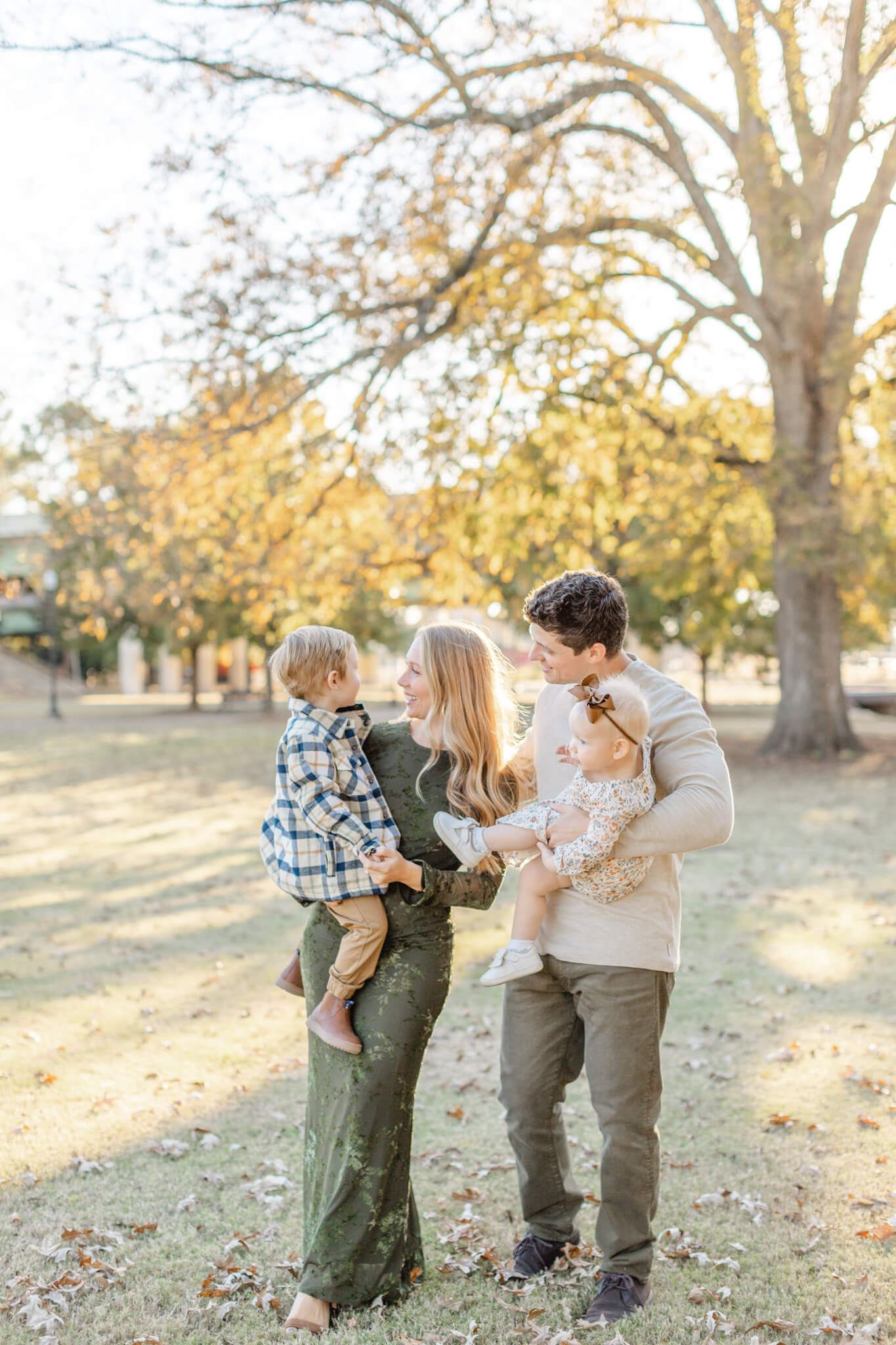 A mom and dad walk in a park lawn carrying their toddler son and daughter in their arms at sunset after finding playgrounds in huntsville al